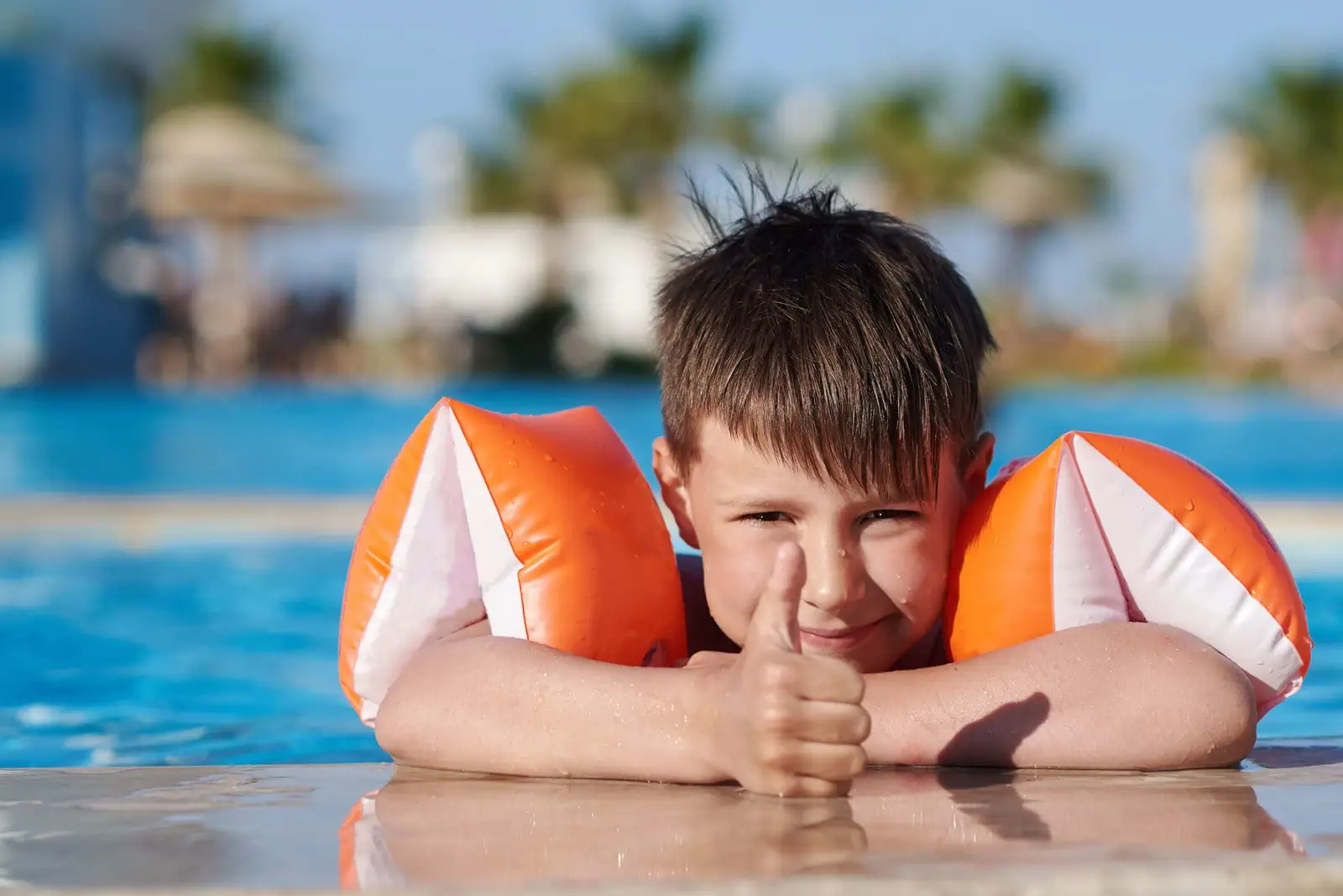 A young boy in pool safety gear giving a thumbs up beside a pool