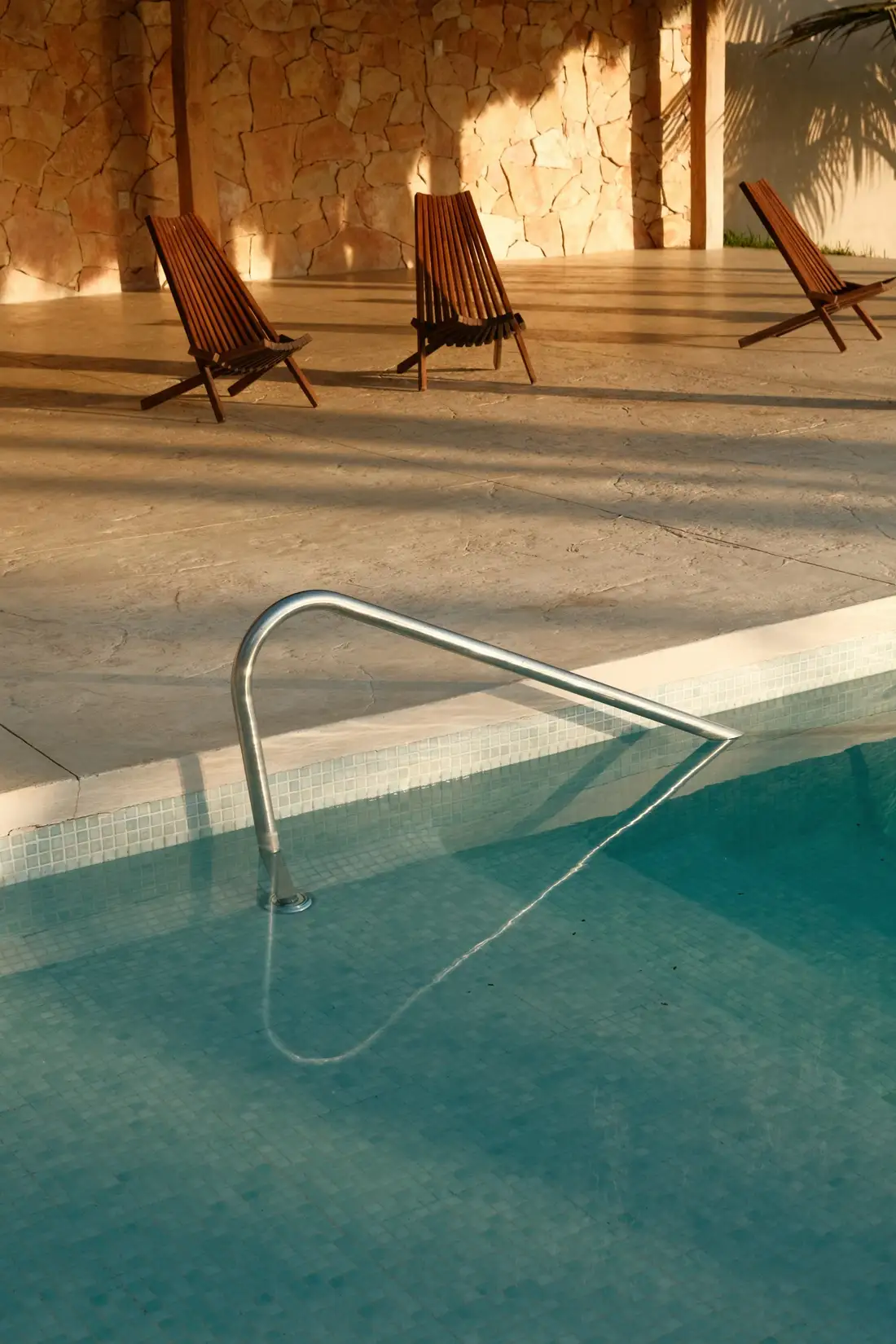 Stainless steel handrail at the edge of a mosaic-tiled swimming pool, with timber slat chairs on sandstone paving behind