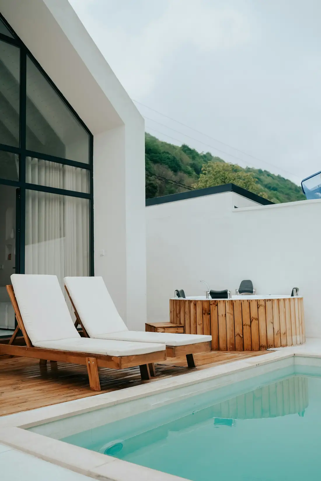 Timber sun loungers and a round cedar spa on the deck of a modern white home beside a swimming pool