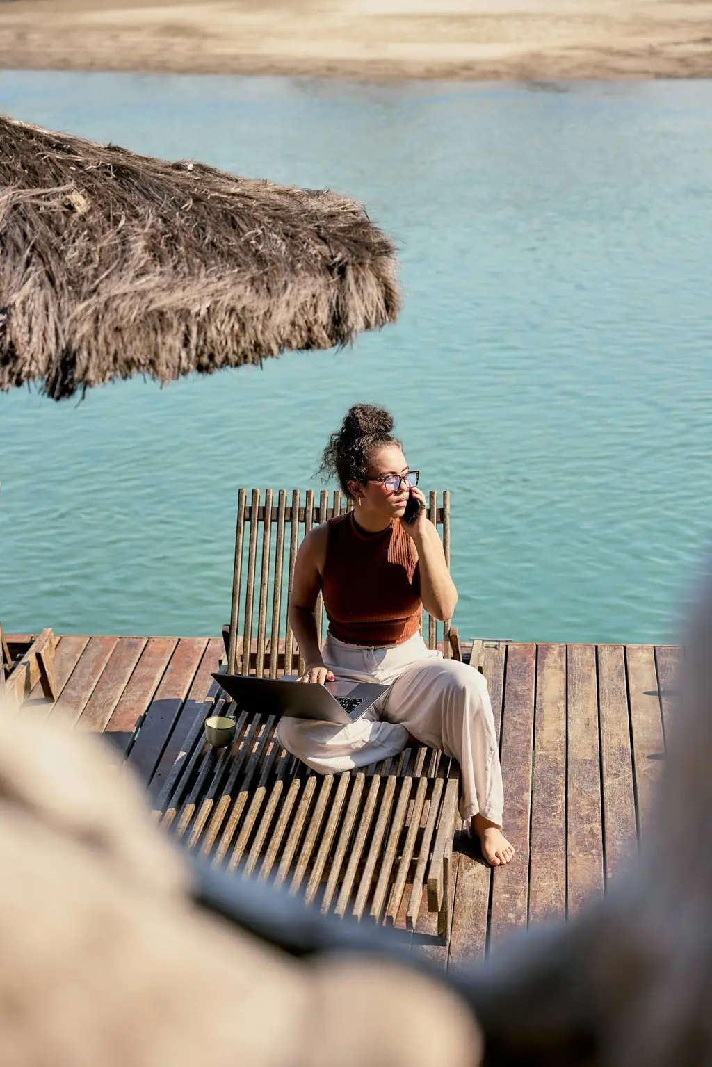 Woman on a phone with a laptop, sitting on a timber deck beside turquoise water